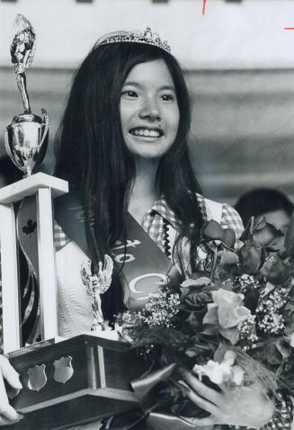 A young smiling Asian woman wearing a tiara holds a bouquet of flowers and a trophy.