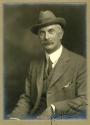 Sepia-toned studio portrait of an adult individual seated and turned slightly to the side, wear…