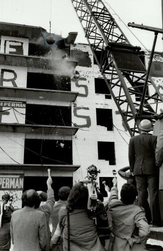 Small group of reporters hold microphones toward multi-level building being demolished by wreck…