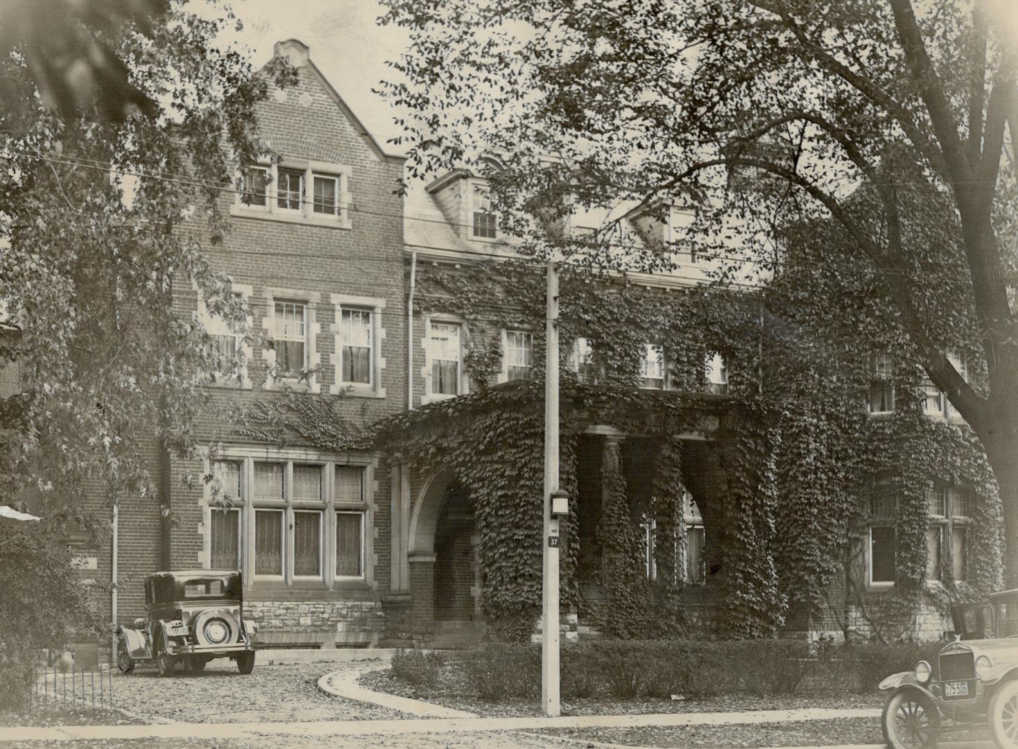 Three-storey, vine covered brick building with roof gables and porte-cochere at entrance.