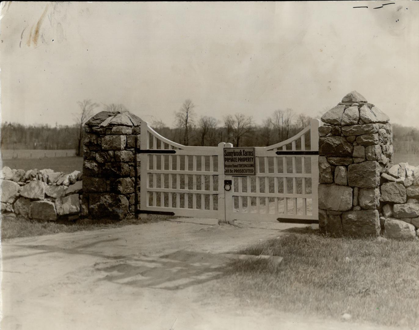 White lattice gate attached to tumbled stone pillars and low stone wall either side of a dirt e ...