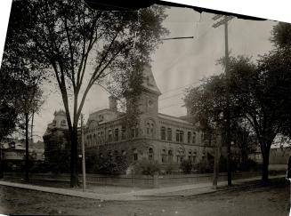 View of treed grounds and perimeter fence enclosing two-storey, vine-covered brick building wit…