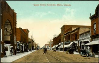 A street scene. Streetcar tracks run along the centre of an unpaved road, used by a mix of comb ...