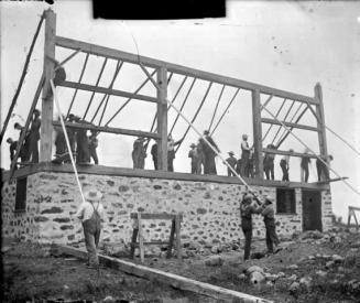 Shows a group of 25 to 30 men, in suspenders and hats, raising a frame for a wall onto a stone  ...