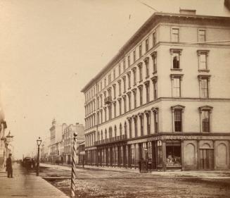 Corner view of a 1860s-era 5-storey hotel. There is a barber pole and a gas streetlight across  ...