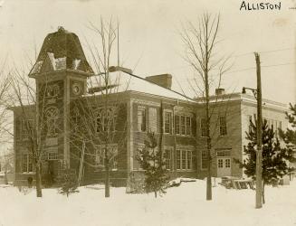 Shows a two-storey brick building with paned sash windows, and a large, ornate tower in the fro…