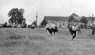 Young women in dark bloomers and white blouses race through a field. Spectators in background.