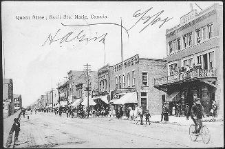 A busy summer street scene, numerous bicycles and horses to down the unpaved street, a streetca ...