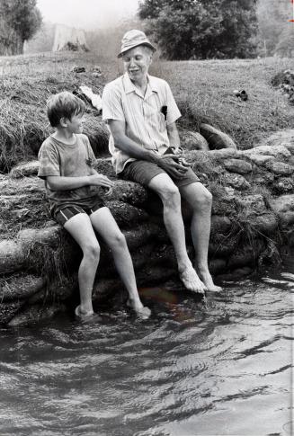 11-year-old Michael Broughton and 60-year-old Roland Adams, who was one of the original campers in 1922, dangle their feet in Cold Creek. Bolton Camp, Ontario