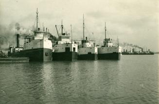 Image shows four ships side by side by the harbour with another ship passing by in the backgrou ...