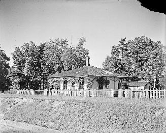 Image shows a cottage within a fenced area with some trees behind it.