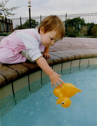 A toddler in a pink outfit leans precariously over the edge of a pool, reaching for a rubber du ...