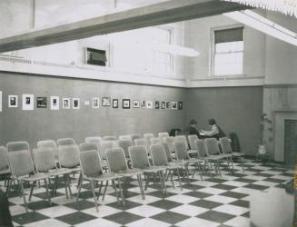 Three rows of empty chairs arranged on a checkerboard pattered floor. A row or artwork hangs on…