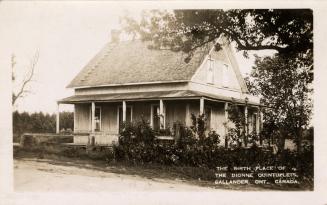 Black and white picture of a wooden house with a porch wrapping around it.