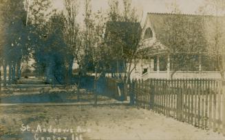 Black and white photograph of a cottage on a sandy street surrounded by a picket fence.