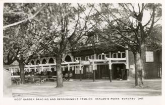 Black and white photograph of a long wooden buildings with tables set up outside the front door ...
