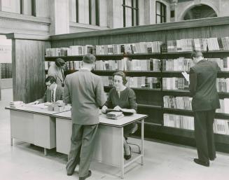 Two librarian, one male and one female, sit at a desk and assist a user while another user brow ...