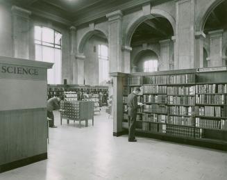 Man standing reading at book shelves in a library with large arches and shelves and a card cata ...