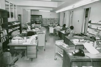 Library office work room with several desk and people seated working and several book trucks an ...