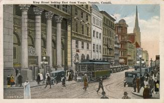 Colorized photograph of a busy street corner with people, cars and street cars.
