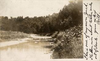 Black and white photograph of a narrow river with trees on the right hand side.
