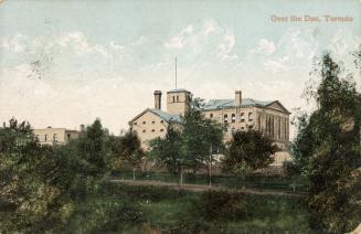 Colorized photograph of a large Victorian jail building as seen in the distance across a river. ...