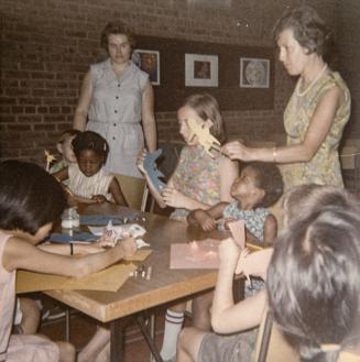 Picture of a group of children making puppets at a table and two women standing helping them.
