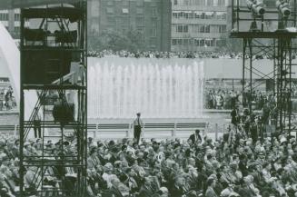 Picture of large crowd seated in public square with fountains in background. 