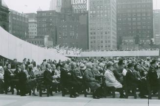 Picture of crowd of people seated in public square with buildings in the background. 