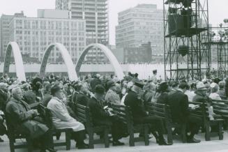 Picture of crowd of people seated in square with fountains and buildings in background. 