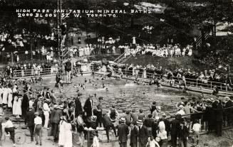 Black and white photograph of crowds of people standing around and swimming in a huge, public p ...