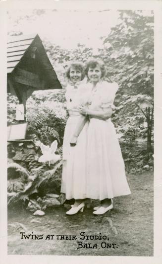 Black and white photograph of two identical women dresses party dresses in a garden.