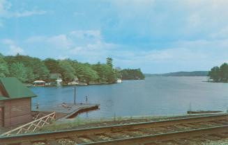Color photograph of railway tracks and a dock with boathouse beside a lake in the summer.