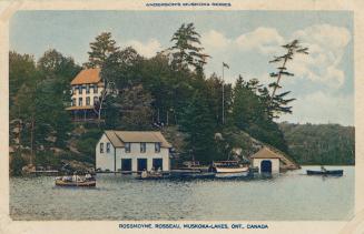Several buildings and boathouses on a rocky ledge in front of a body of water with people in bo ...