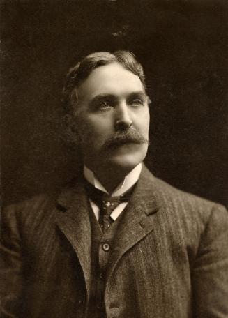 A formal photograph of a middle-aged man who is wearing a suit and tie. He has short hair and a ...