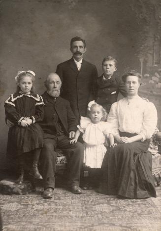 A formal, posed photograph of a family taken in a studio, with three adults and three children. ...