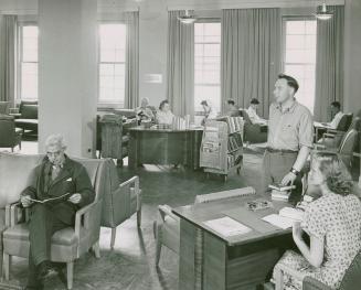 Men sit reading or standing in a library and two librarians sit at desks. 