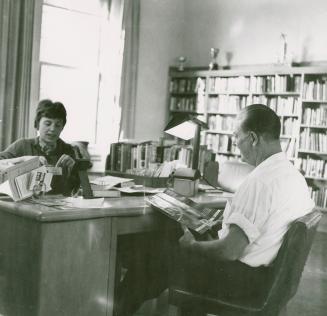 A librarian sits at a desk speaking with a man seated in front of her. 