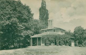 Black and white photograph of a two story home with a porch wrapping around it in a wooded area…