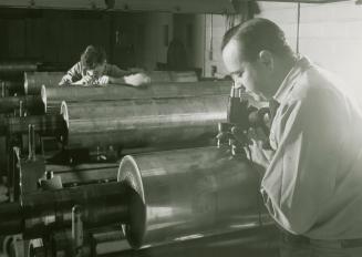 A photograph of two men using microscopic devices to examine images on cylinders during the pro ...