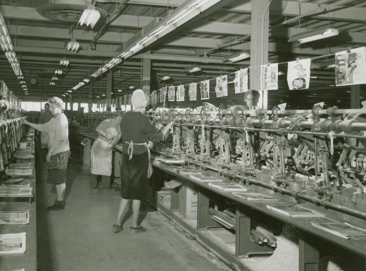 A photograph of men and women in front of a metal assembly line, with piles of pages of catalog ...