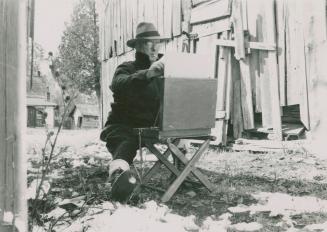 A photograph of a man sitting on a small chair and painting on a canvas, which is propped up in ...