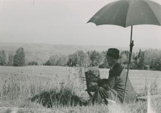 A photograph of a man sitting on the ground in a grassy meadow underneath an umbrella, painting ...
