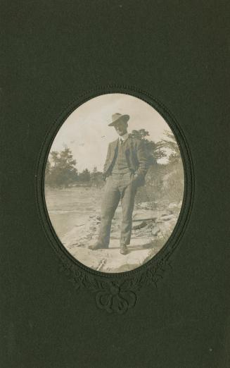 A photograph of a man standing and posing for a photograph beside a river. He is wearing a hat  ...