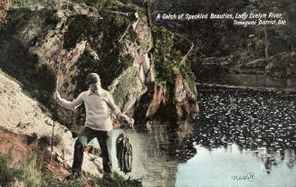 Colorized photograph of a man holding a large catch of fish in a rocky area beside a lake.