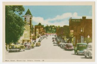 Colorized photograph of the main street in a small town. Cars are parked bumper to bumper on ea ...