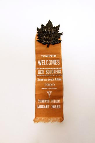 A metal pin in the shape of a maple leaf attached to an ochre ribbon. The ribbon reads "Toronto ...