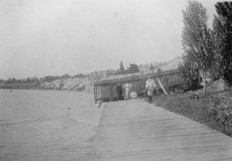 A photograph of a boardwalk beside a body of water. There are three people walking on the board ...