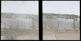 A stereo photograph of a harbour, with ships docked on both sides of a body of water. Buildings…