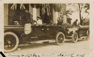 A photograph of two antique cars parked in front of a large building or hall. There are British…